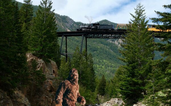 Train crossing bridge with Sasquatch walking across forest